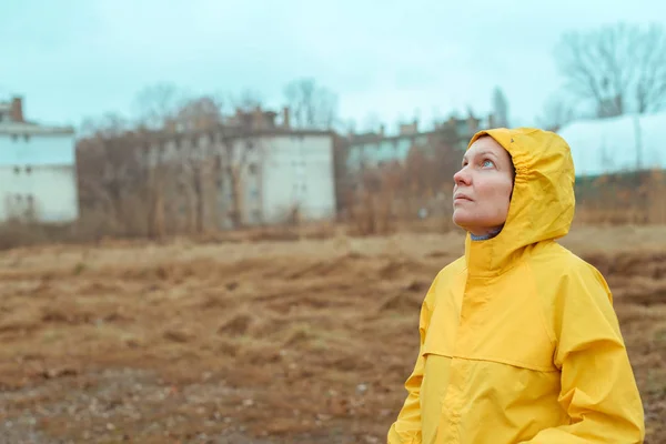Woman in yellow raincoat looking up at rainy clouds