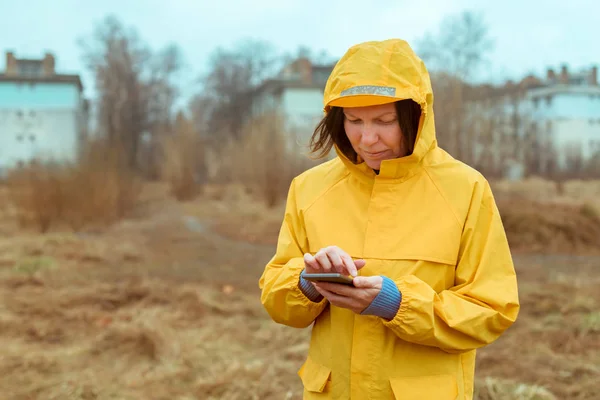 Mulher em capa de chuva amarela mensagens de texto no celular ao ar livre — Fotografia de Stock