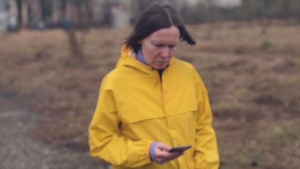 雨の日に草原を歩くと 陰鬱な午後 スローモーションのハンドヘルド映像に心配と悲しい女性の携帯電話でメッセージを読んでレインコートの女 — ストック動画
