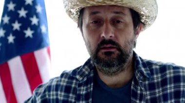 Kamera ve konuşurken, yetişkin erkek kişinin portre hasır şapka ve arka planda ABD bayrağı ile ekose gömlek arıyorum ciddi Amerikan çiftçi