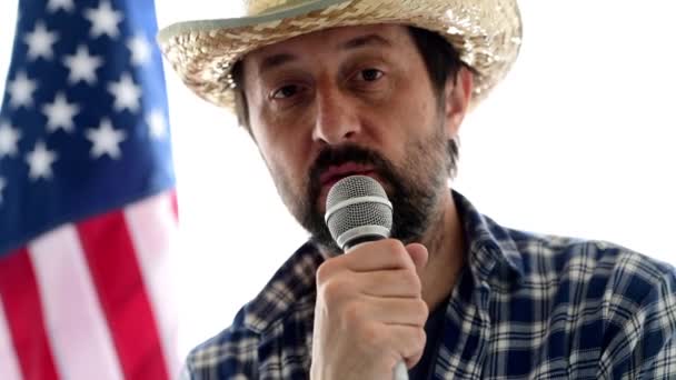 麦わら帽子とバック グラウンドでアメリカ国旗の格子縞のシャツでカメラとマイクで話している 大人の男性人の肖像画を見て深刻なアメリカの農家 — ストック動画