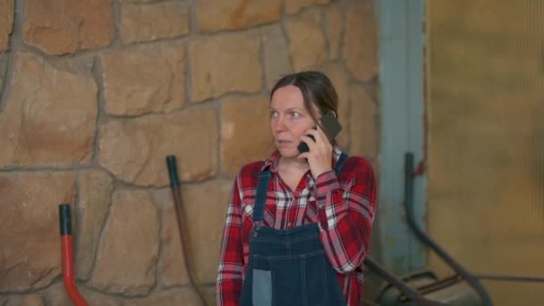 忧心忡忡的女农场主在马厩里用手机说话 穿着格子呢衬衫的成年妇女和电话交谈中的全裤牛仔 — 图库视频影像
