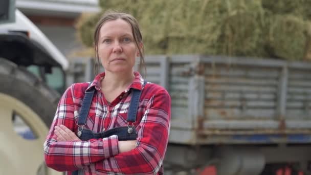 女农民在干草车前摆姿势 女农场工人穿着格子衬衫和围兜工作服的画像 拖拉机拖车上装满了奶牛场牲畜饲料干草捆 — 图库视频影像