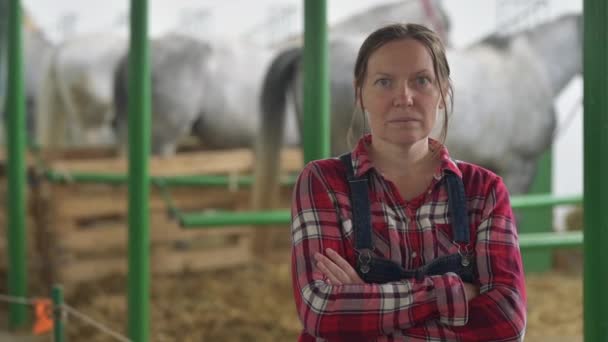 カメラを見て馬小屋の女性牧場の肖像画 農場労働者としてチェック柄のシャツとジーンズビブのオーバーオールを着ている大人の女性 — ストック動画