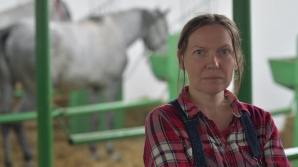 女牧场主的肖像在马稳定看相机 成年妇女穿着格子衬衫和牛仔裤围兜工作服作为农场工人 — 图库视频影像
