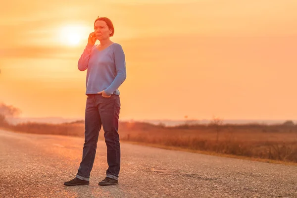 Mulher de pé na estrada rural e falando no telefone móvel — Fotografia de Stock