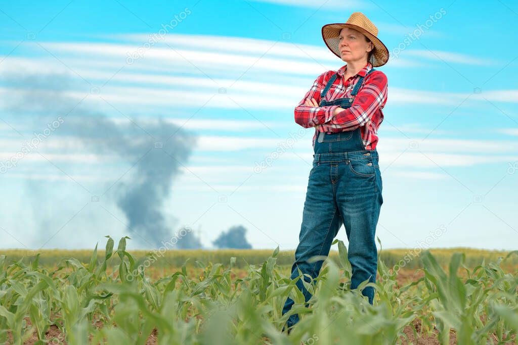 Concerned female farmer in corn field looking at black smoke on horizon, concept of insurance in agriculture and farming