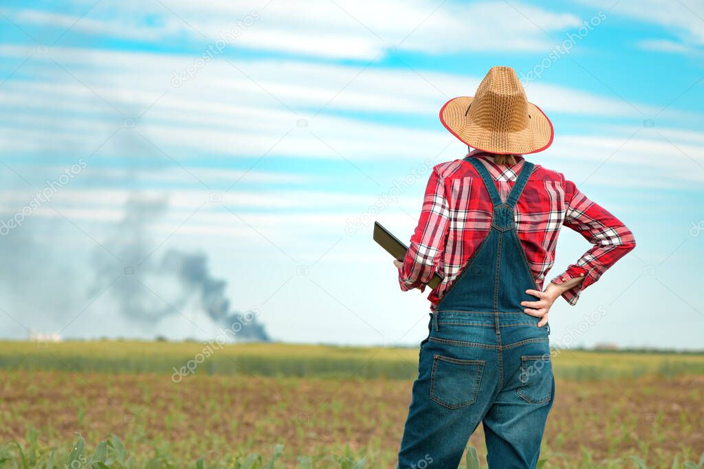 Concerned female farmer with digital tablet in corn field looking at black smoke on horizon, concept of insurance in agriculture and farming