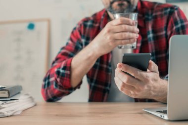 Gig işçisi su içiyor ve ev ofisinde cep telefonu kullanıyor serbest proje çalışmaları sırasında.