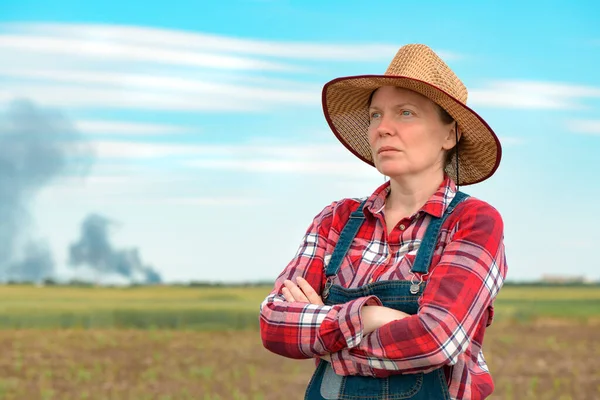 Concerned female farmer in corn field looking at black smoke on horizon, concept of insurance in agriculture and farming