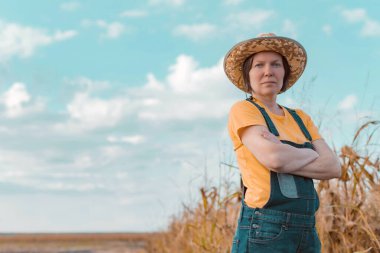 Female corn farmer looking over cornfield, portrait of woman agronomist with straw hat and jeans bib overalls clipart