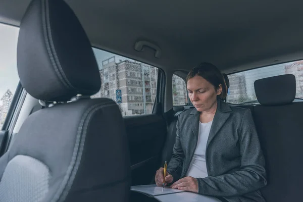 Businesswoman doing business paperwork on car back seat, adult caucasian female businessperson executive analyzing business results while traveling by automobile