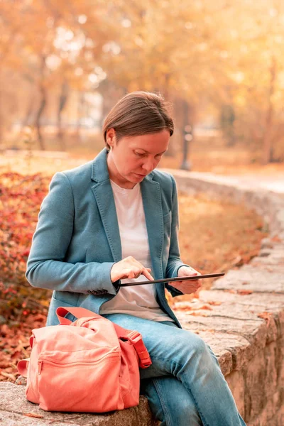Woman using digital tablet on park bench in autumn during working hours break, selective focus