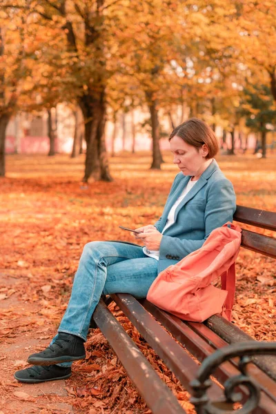 秋の晴れた日に公園のベンチに座っている間 休憩中に携帯電話を使用している女性 — ストック写真