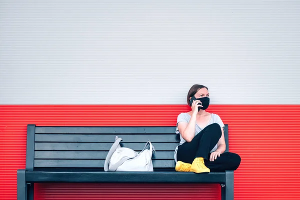 戴着防护面罩的女人坐在长椅上 一边在街上用手机说话 一边又带着新的正常观念 — 图库照片