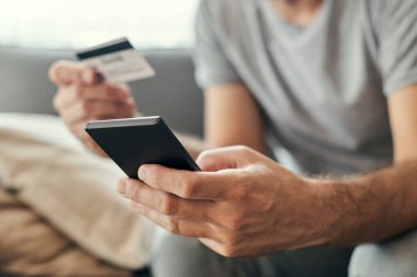Cep telefonu ve kredi kartıyla online alışveriş yapan bir adam oturma odasındaki kanepesinden internetten alışveriş yapan yetişkin bir erkek.