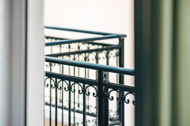 Zarif, siyah işlemeli demir balkon korkulukları açık bir pencereden görülüyor. Yumuşak odaklı, mimari detaylar ve sakin bir şehir hayatı hissi veriyor. Seçici odak.