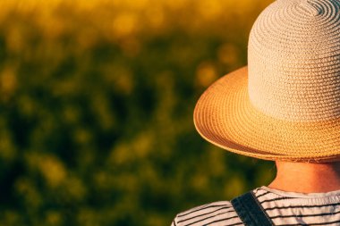 Hasır şapkalı kadın çiftçinin dikiz görüntüsü çiçek açan kolza tohumu tarlasında duruyor ve ufukta mesafeye bakıyor, seçici bir odak noktası.