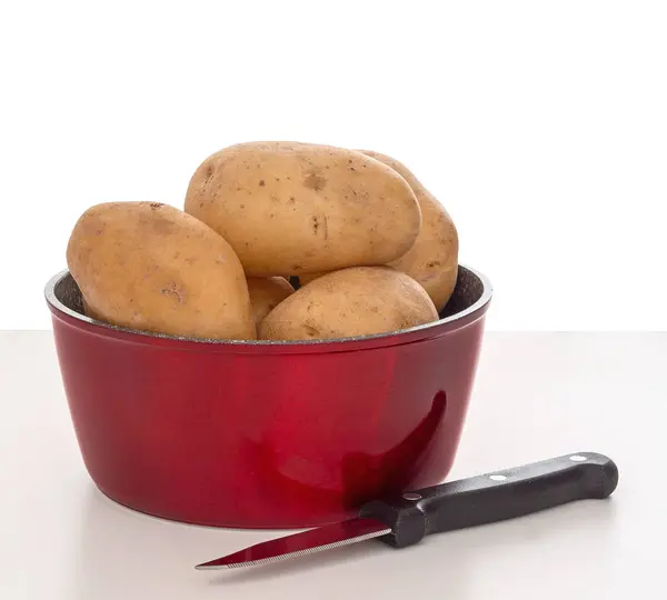 食事の準備。ナイフ、白い背景を持つ光沢のある赤い鍋で生のジャガイモ. — ストック写真