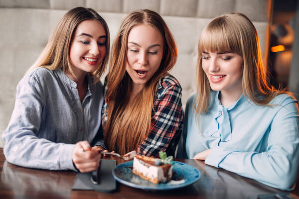 three smiling young women eating sweet cake in cafe