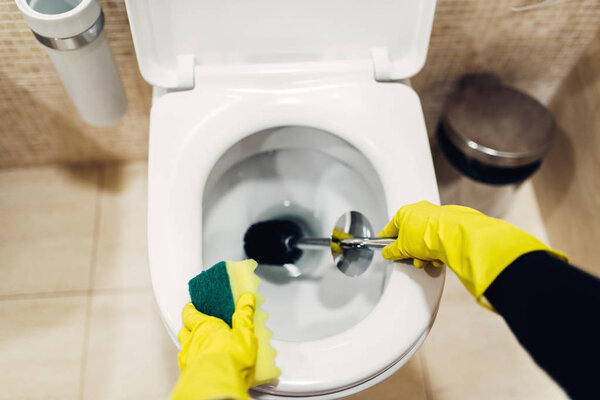maid in rubber gloves cleaning toilet with brush, hotel restroom interior on background. Professional housekeeping service, sanitary processing
