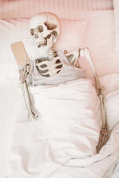 人的骨骼用手提电话在手躺在床上 滑稽笑话 — 图库照片