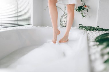 Female legs dips into the bath with foam. Bathroom interior with window on background