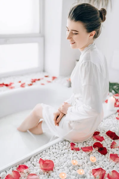 白いバスローブとタオル頭お風呂の端に座っているに魅力的な女性は バラの花びらで飾られました 豪華なバスルームのインテリア — ストック写真