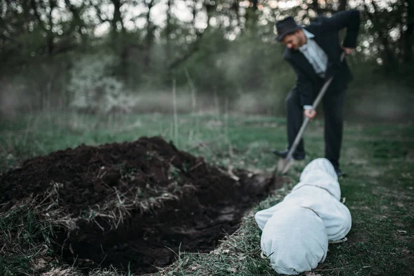 Killer is digging a grave for the victim in forest, the body wrapped in a canvas, serial maniac concept