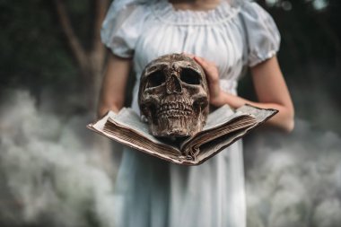 Beyaz elbiseli kadın kurban kitap ve insan kafatası el, orman arka plan üzerinde tutar. Fotoğraf korku tarzı, şeytan çıkarma
