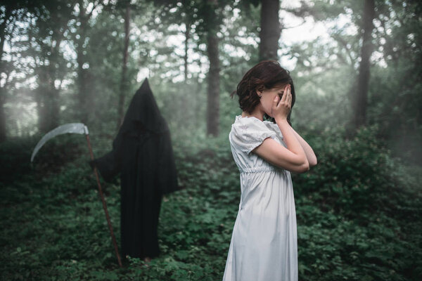 Young female victim in white dress in the forest, death in a black hoodie and with a scythe on background. Photo in horror style, exorcism