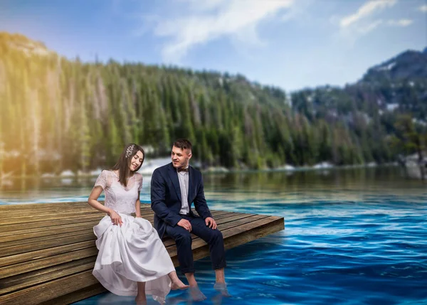 新娘和新郎坐在码头上 在水中湿润他们的脚 在湖上浪漫的场景 山和森林的背景 夏季自然景观 蜜月或婚礼旅行 — 图库照片