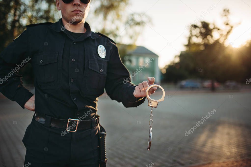 Serious police officer in uniform and sunglasses holds handcuffs, front view. Cop at the work. Law protection concept, safety control job