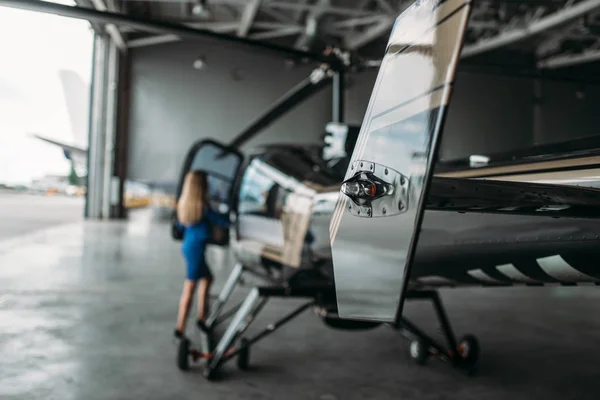 Female flight attendant stands against helicopter in hangar with opened gates. Stewardess in uniform near copter. Private air transportation