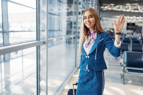 Sexy stewardess with suitcase wave her hands in airport waiting room. Air hostess in suit with luggage, flight attendant occupation