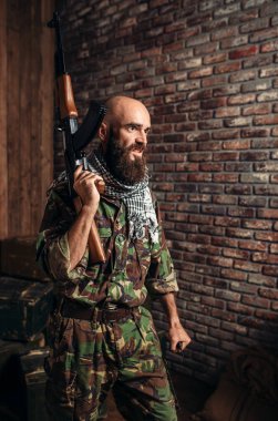 Üniformalı terörist Kalaşnikof tüfeği elinde silah ile erkek mojahed tutar. Terörizm ve terör, asker haki kamuflaj, Yakýt veya kimyasallarýn arka plan üzerinde varil