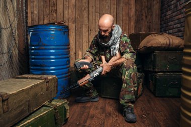 Üniformalı terörist dergi Kalaşnikof tüfek ekler. Terörizm ve terör, varil yakıt veya kimyasal maddeler ve kutu mermi arka plan üzerinde silah, kamuflaj asker şarj eder