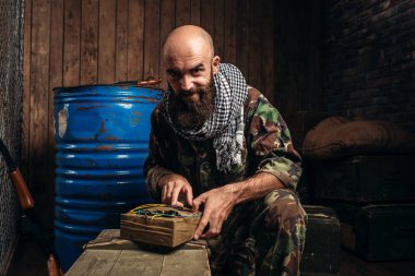 Sakallı terörist yapımı bir bomba, patlayıcı ile erkek mojahed üniformalı. Terörizm ve terör, haki kamuflaj, asker yakıt veya kimyasal maddeler ve kutu mermi arka plan üzerinde varil