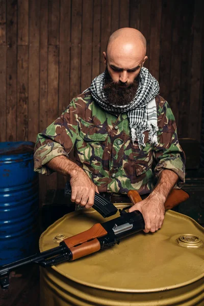恐怖分子在制服把杂志插入卡拉什尼科夫步枪 恐怖主义和恐怖 战士在伪装充电武器 桶燃料或化学品和箱子弹药在背景上 — 图库照片