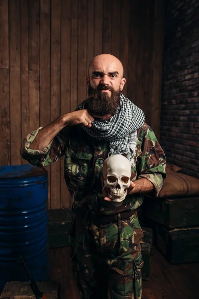 制服のテロリストは Mojahed の手で人間の頭蓋骨を保持しています テロとテロの恐怖に カーキ色の迷彩 バレルの燃料や化学薬品の背景にひげを生やした兵士 — ストック写真