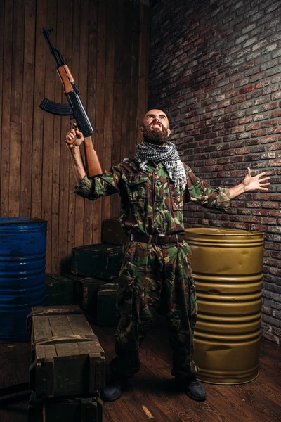 ひげを生やしたテロリスト カラシニコフ銃を保持しています テロとテロ カーキ色の迷彩 軍用ヘリコプターと背景にタンクの兵士 — ストック写真