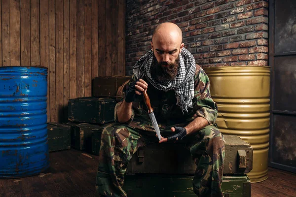制服のテロリストの弾薬箱の上に座って手にナイフ テロとテロ 武器の工廠 バレルの燃料や化学薬品の背景に迷彩の兵士 — ストック写真