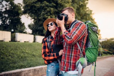 Hikers with backpacks go sightseeing in tourist town and makes photo for memory. Summer hiking. Hike adventure of young man and woman clipart