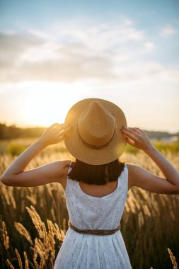 Kadın beyaz elbise ve hasır şapka buğday alanında günbatımı üzerinde yürüme. Yaz çayır üzerinde kadın kişi