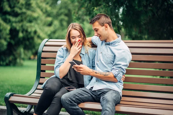 Phone addiction, smiling couple using gadget on the bench in summer park. Social addicted people