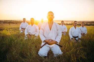 Karate group sitting on the ground and meditates, training in summer field. Martial art school on workout outdoor, relaxation in nature clipart