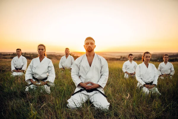 Karate group sitting on the ground and meditates, training in summer field. Martial art school on workout outdoor, relaxation in nature