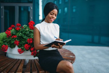 Bankta oturan işkadını ve ofis binası önündeki Defteri'nde yazıyor. Beyaz bluz siyah iş kadını gülümseyen açık havada çalışır