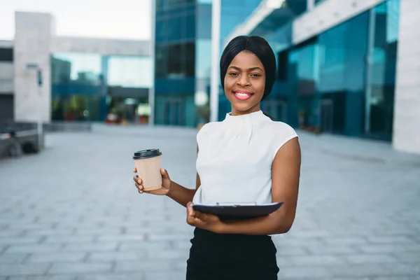 Business woman with cardboard coffee cup and notepad outdoors, office building on background. Black businesswoman in skirt and white blouse