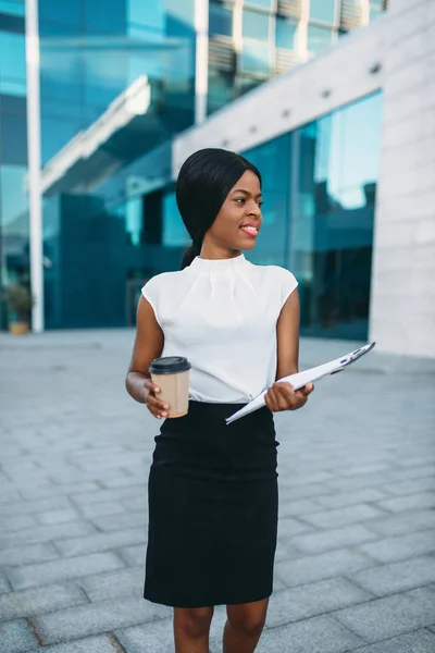 Business woman with cardboard coffee cup and notepad outdoors, office building on background. Black businesswoman in skirt and white blouse
